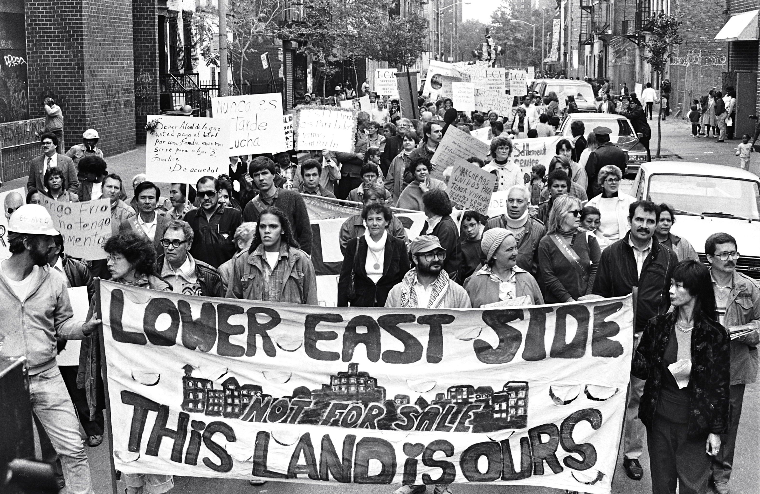 A black and white photo of a group of adults marching down a Lower East Side street. The people in the front hold a banner that says: "Lower East Side. This Land is Ours."